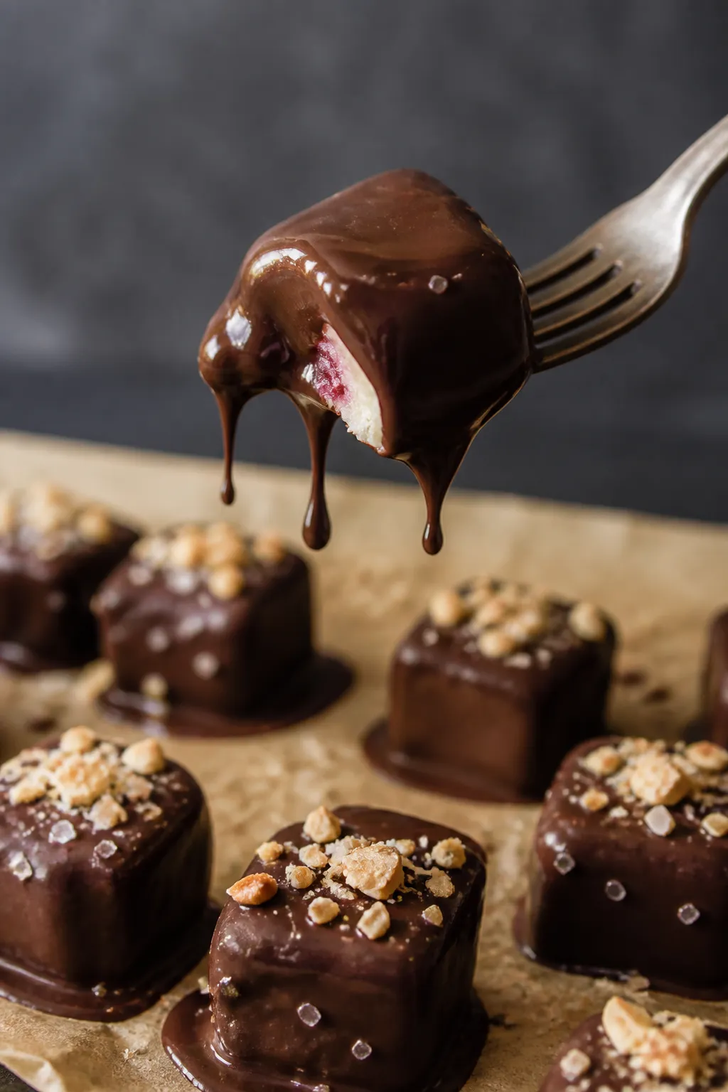 Close-up detail shot: A chilled cheesecake bite mid-dip in glossy melted sugar-free dark chocolate, 